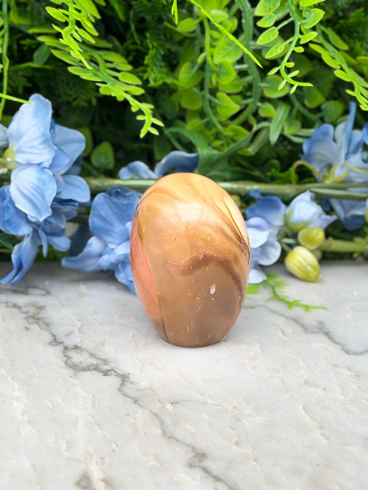 a brown and orange stone, possibly a jade or amber, placed on a marble surface surrounded by blue flowers.