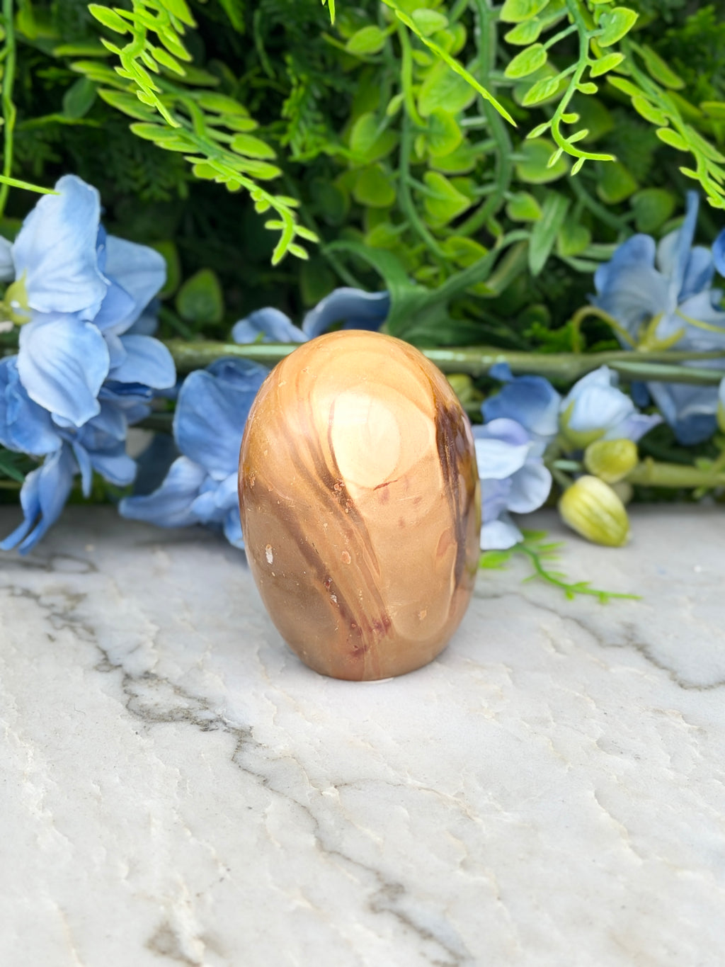 a wooden egg-shaped object with a natural wood grain texture, placed on a marble surface. The background includes a variety of green plants and blue flowers, creating a visually appealing and harmonious composition.