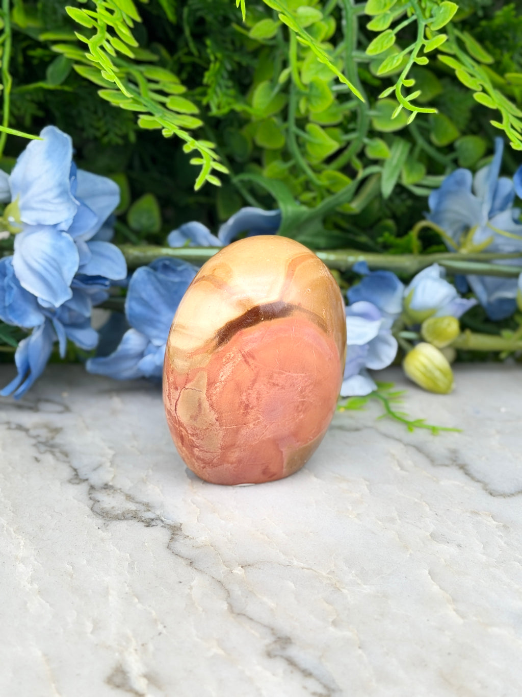 a pinkish-brown stone or gemstone, surrounded by blue flowers and green foliage, creating a visually appealing and harmonious composition.