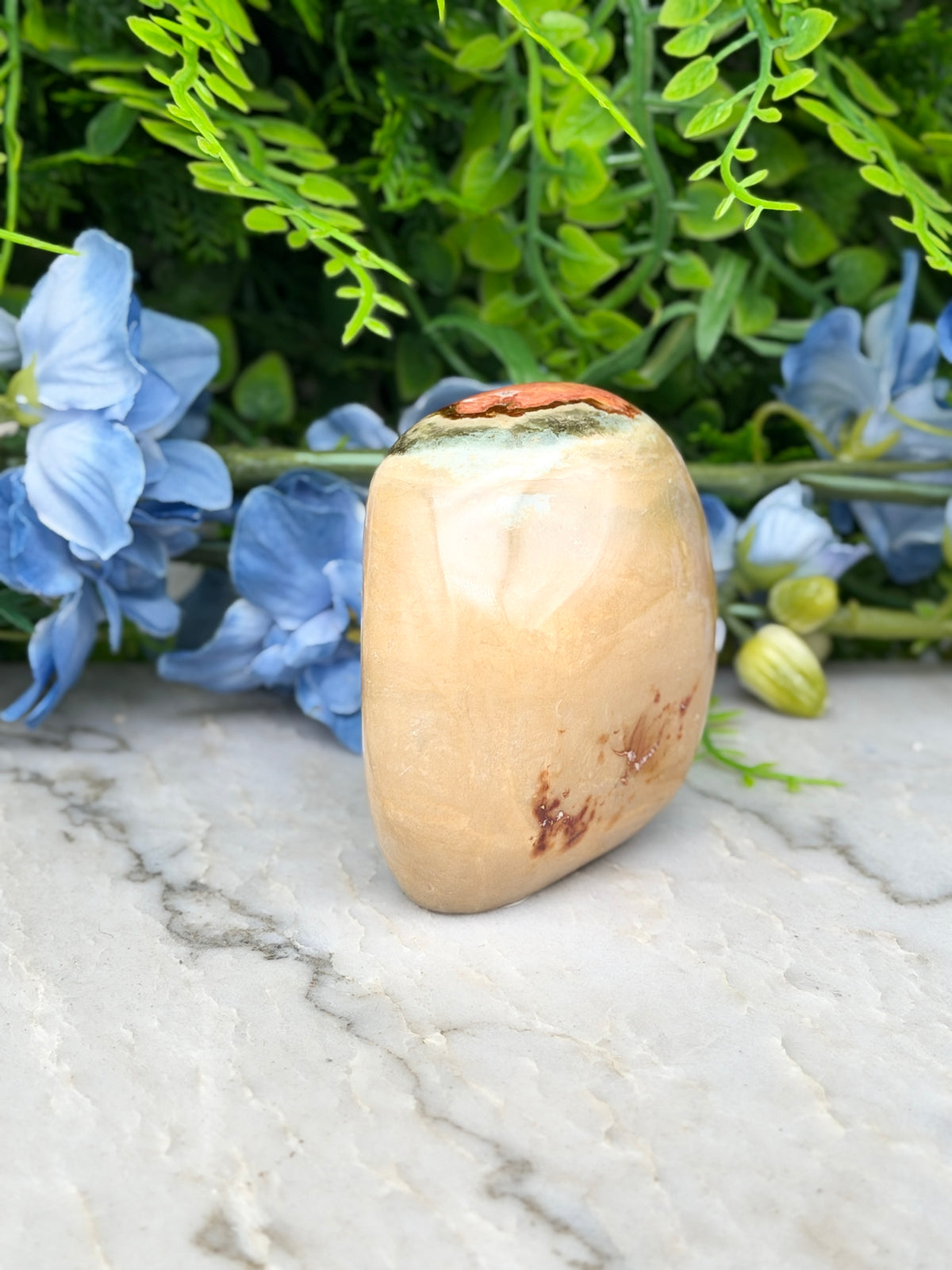 a tan-colored stone or rock with a brownish hue, placed on a marble surface. The stone is surrounded by a variety of blue flowers, creating a visually appealing contrast.