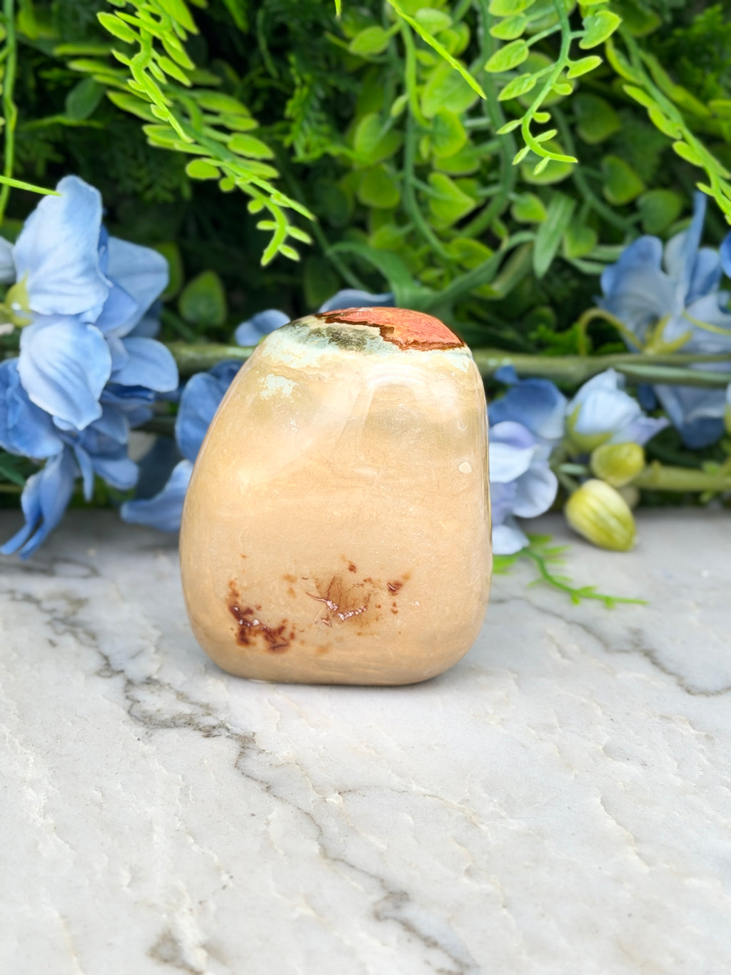 a light brown stone with a small orange spot on it, placed on a marble surface. The stone is surrounded by blue flowers and green foliage, creating a natural and serene setting.