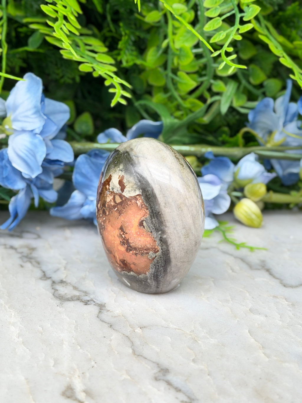 a natural stone, possibly an amethyst, with a unique pattern of orange and gray hues, set against a backdrop of lush green foliage and delicate blue flowers.