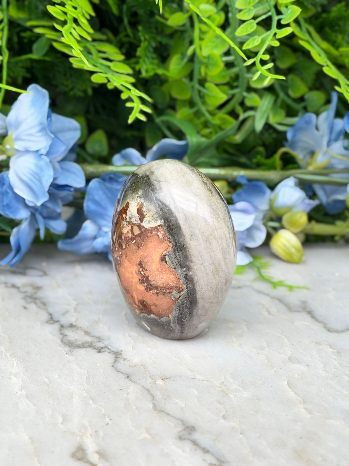 a natural stone, possibly an amethyst, with a unique pattern of orange and gray hues, set against a backdrop of lush green foliage and delicate blue flowers.