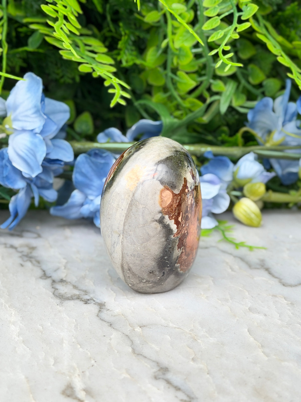a large, oval-shaped stone with a mix of brown and gray hues, placed on a marble surface. The stone is surrounded by a variety of blue flowers, creating a visually appealing contrast.