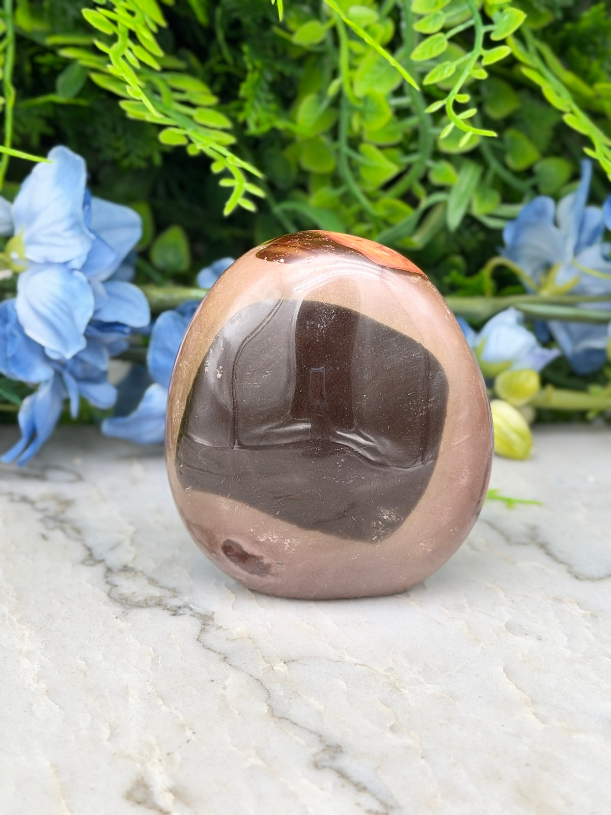A brown and black stone sits on a marble surface, surrounded by blue flowers and green foliage.