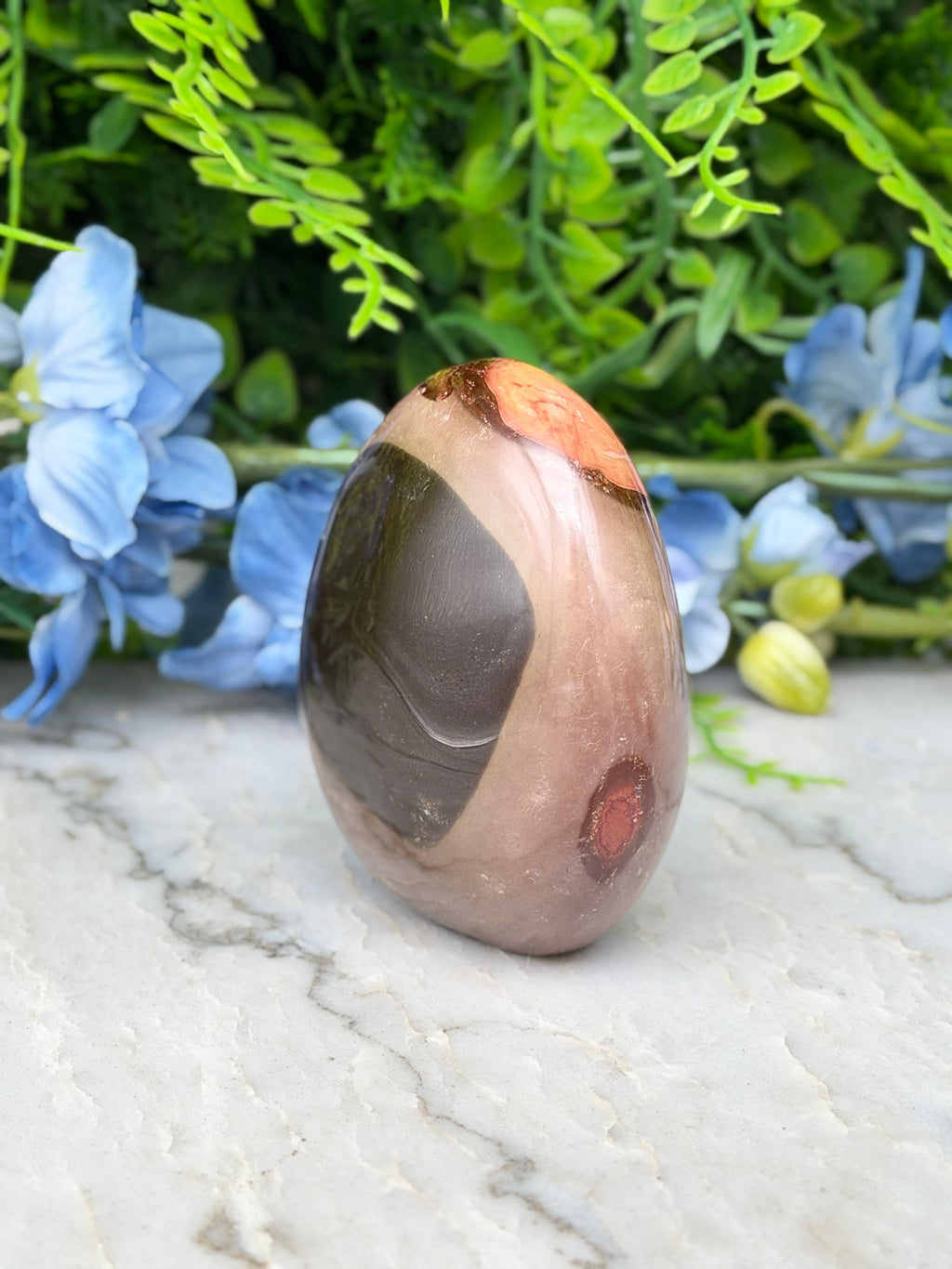 a brown and orange stone with a black and brown pattern, placed on a white marble surface. The stone is surrounded by blue flowers, creating a visually appealing contrast.