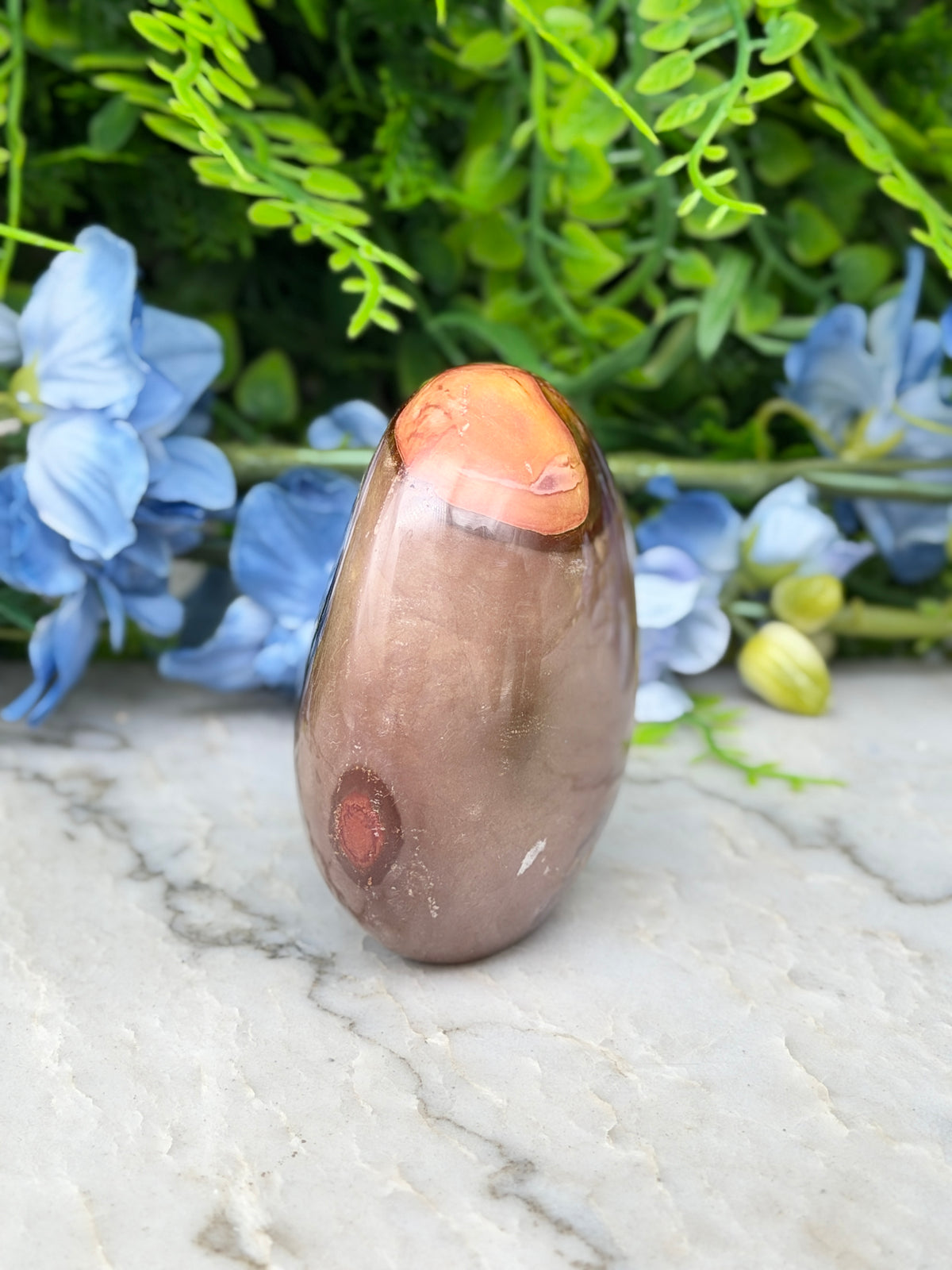 a brown, oval-shaped stone with a red spot on it, placed on a marble surface. The stone is surrounded by blue flowers, creating a visually appealing contrast.
