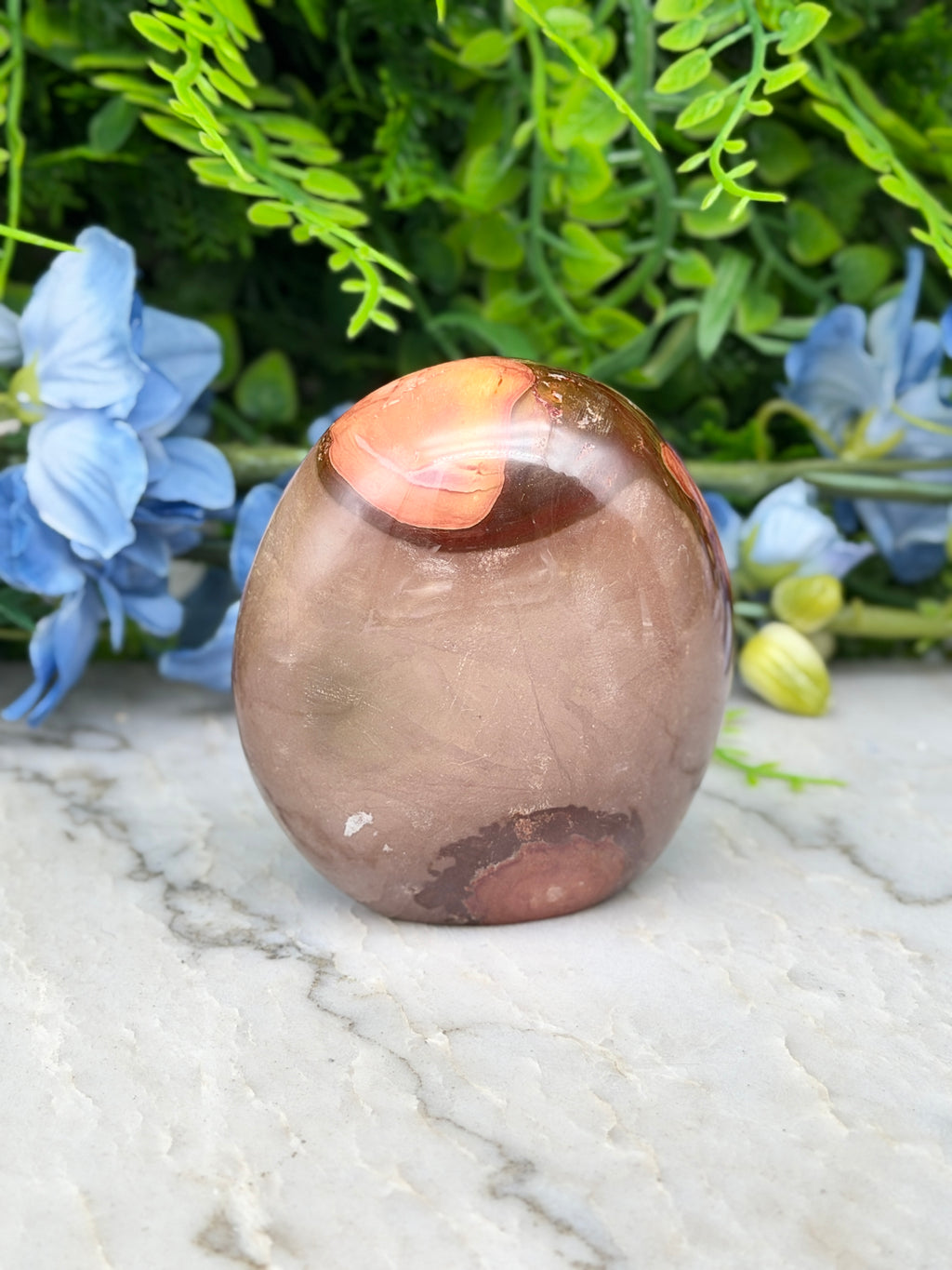 a brown, oval-shaped stone with a reddish-brown hue, placed on a white marble surface. The stone is surrounded by a variety of green foliage, creating a natural and serene setting.