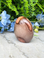 a brown, oval-shaped stone with a reddish-brown hue, placed on a white marble surface. The stone is surrounded by a variety of blue flowers, creating a visually appealing contrast.