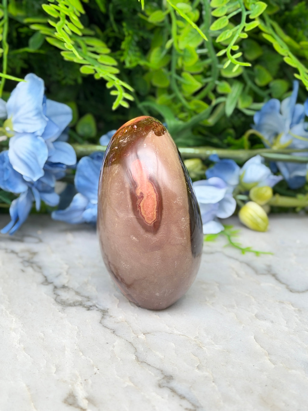 a brown, oval-shaped rock with a reddish-brown hue, placed on a white marble surface. The rock is surrounded by a variety of blue flowers, creating a visually appealing contrast.