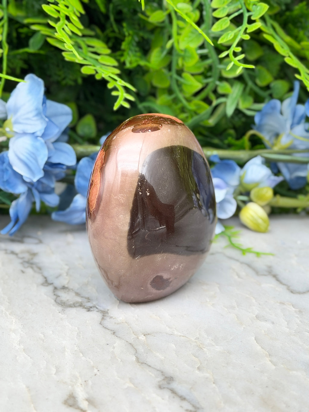 a brown, oval-shaped stone or gemstone placed on a marble surface, surrounded by blue flowers.