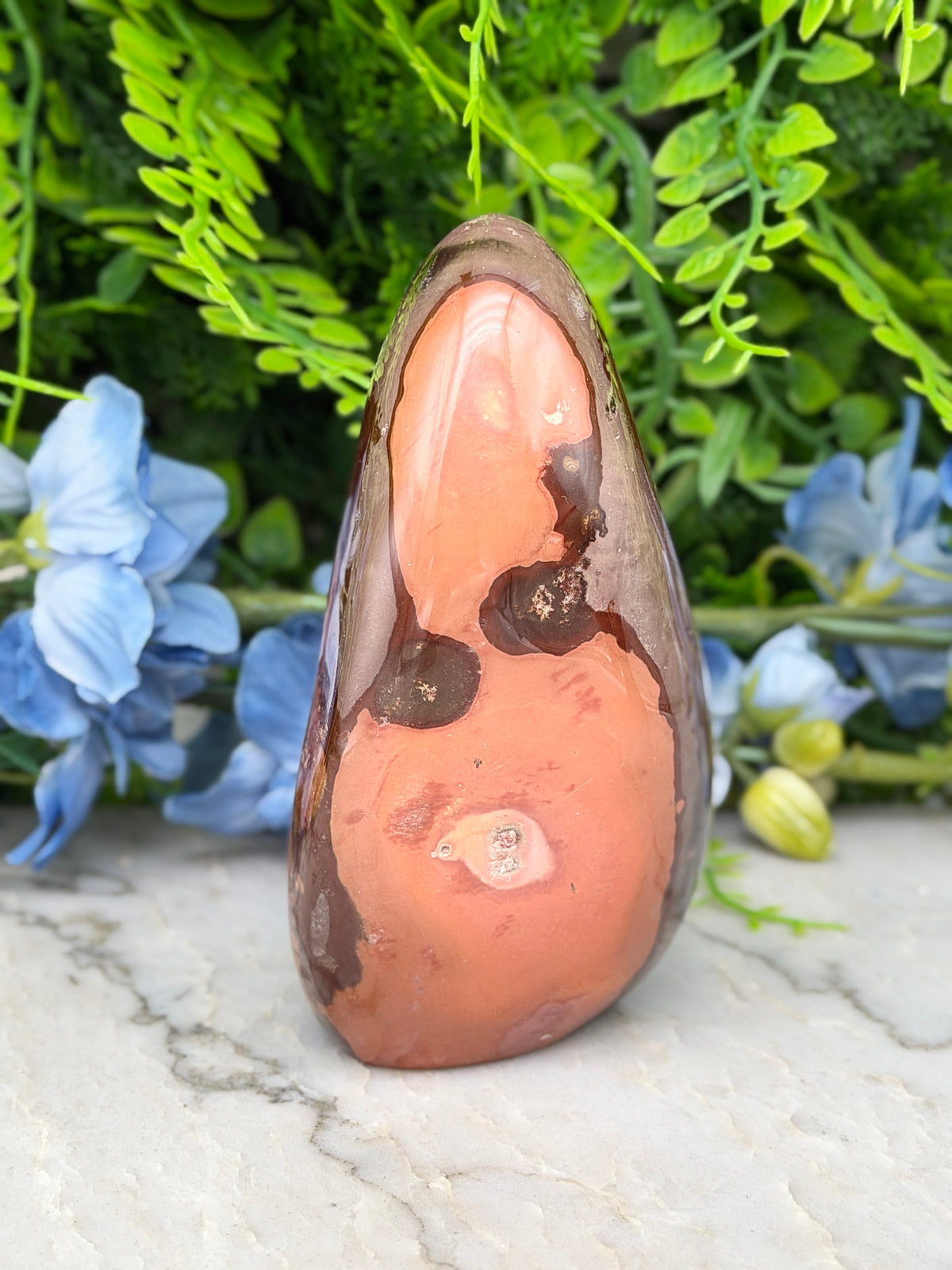 a unique, irregularly shaped rock with a reddish-brown hue, set against a backdrop of lush green foliage and delicate blue flowers.