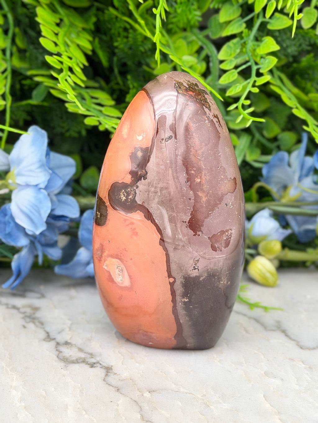 a large, irregularly shaped rock with a mix of orange and brown hues, set against a backdrop of lush green foliage and delicate blue flowers.
