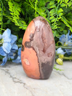 a large, irregularly shaped rock with a mix of orange and brown hues, set against a backdrop of lush green foliage and delicate blue flowers.