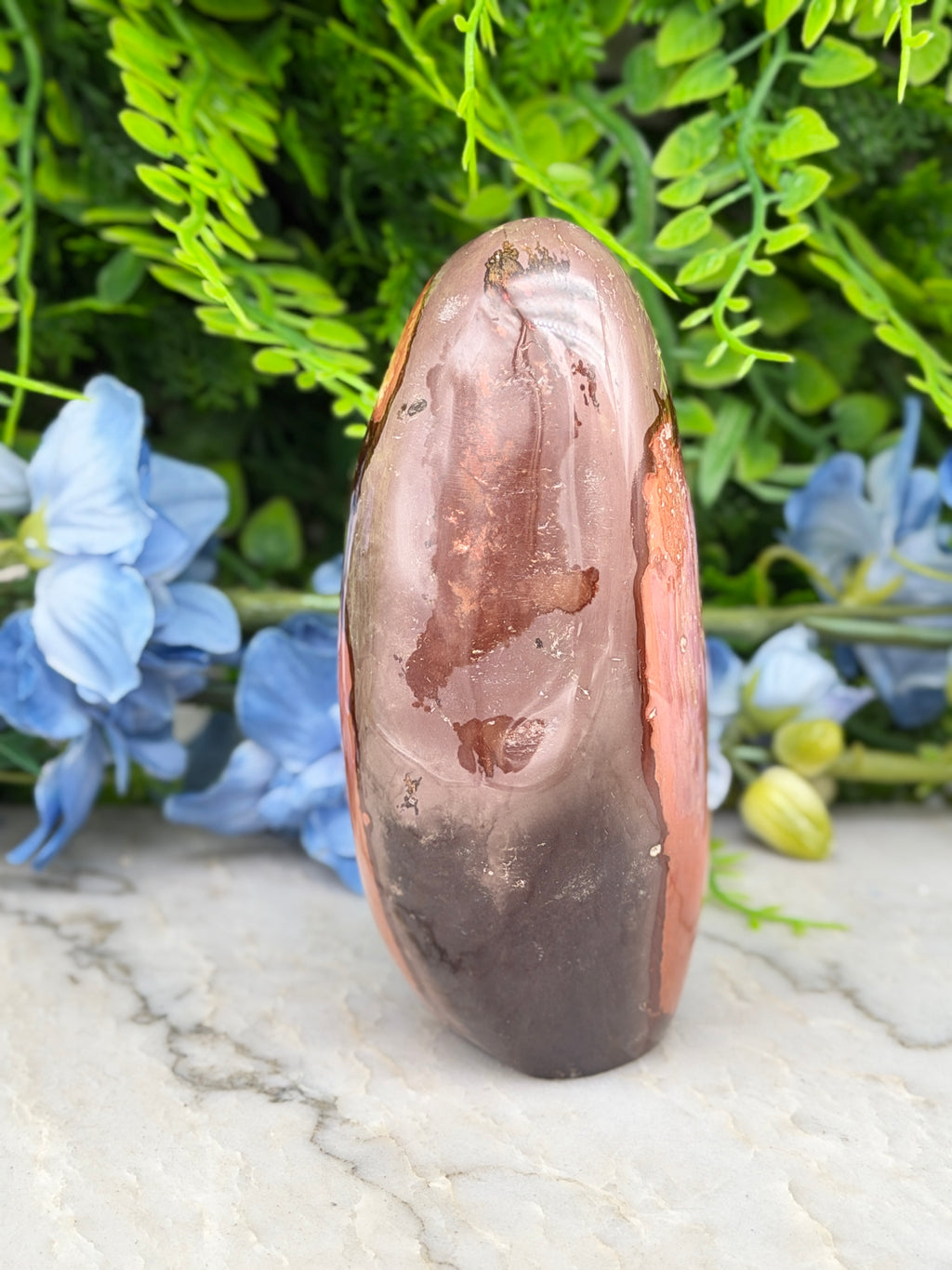 a brown, oval-shaped rock with a reddish-brown hue, placed on a white surface. The rock is surrounded by a variety of green plants and blue flowers, creating a visually appealing and natural setting.