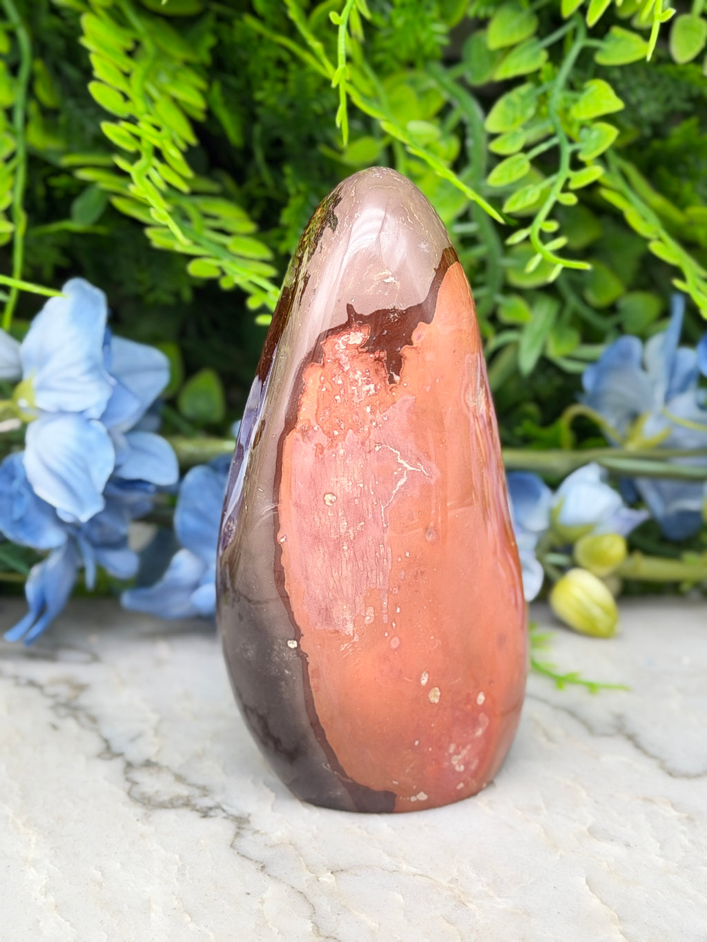 a unique, multi-colored rock formation with a striking combination of orange, brown, and white hues, set against a backdrop of lush green foliage and delicate blue flowers.