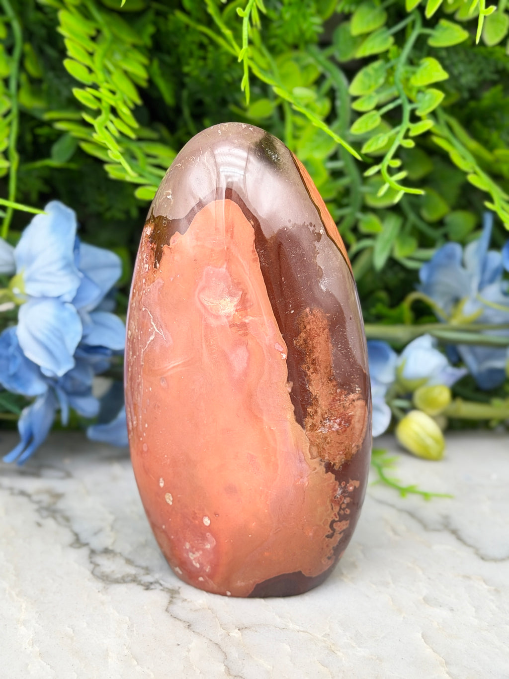 a large, irregularly shaped rock with a mix of orange and brown hues, set against a backdrop of lush green foliage and delicate blue flowers.
