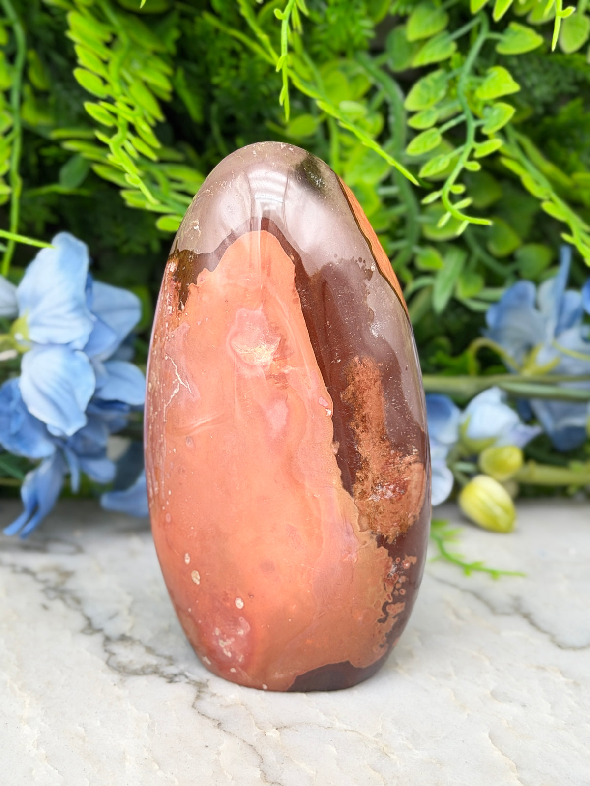 a large, irregularly shaped rock with a mix of orange and brown hues, set against a backdrop of lush green foliage and delicate blue flowers.