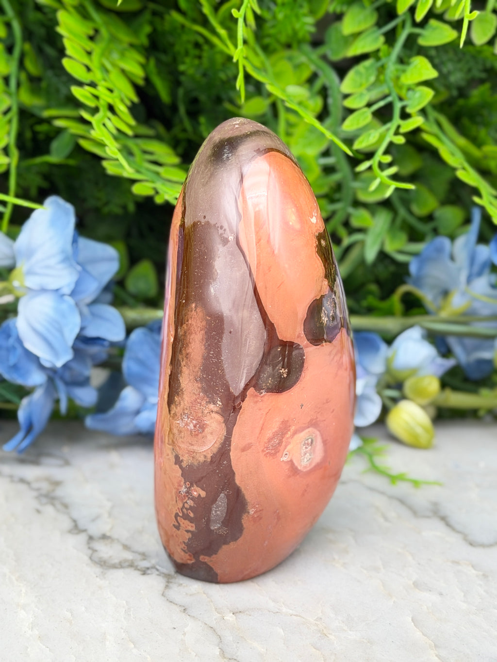 a jadeite, a type of jadeite gemstone, with a unique orange and brown color pattern. The jadeite is placed on a white surface, surrounded by blue flowers and green foliage.