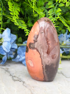 a large, irregularly shaped rock with a mix of orange and brown hues, set against a backdrop of lush green foliage and delicate blue flowers.