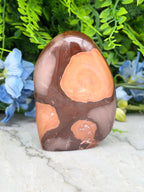 A brown and orange jadeite gemstone is displayed on a white marble surface, surrounded by blue flowers.