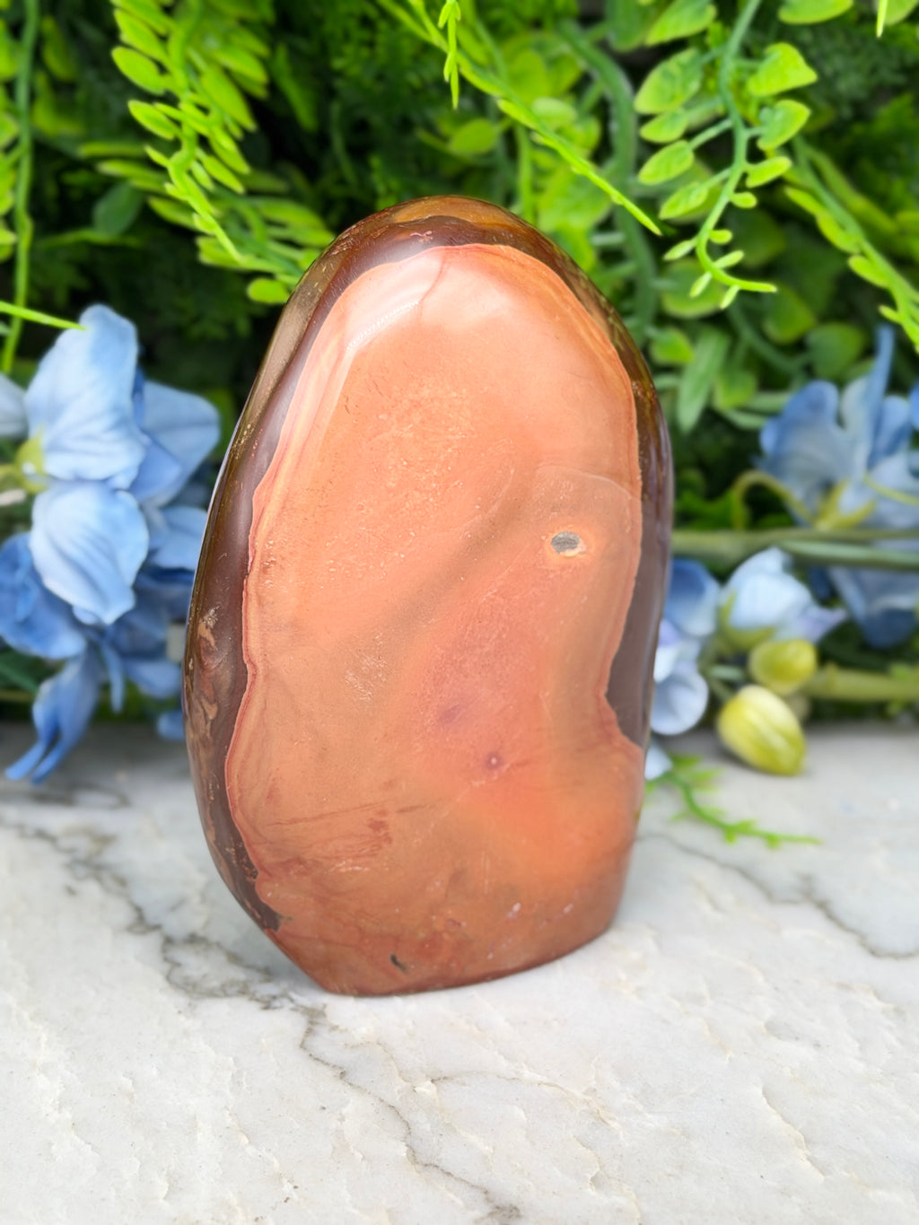 a large, irregularly shaped rock with a reddish-brown hue, set against a backdrop of lush green foliage and delicate blue flowers.