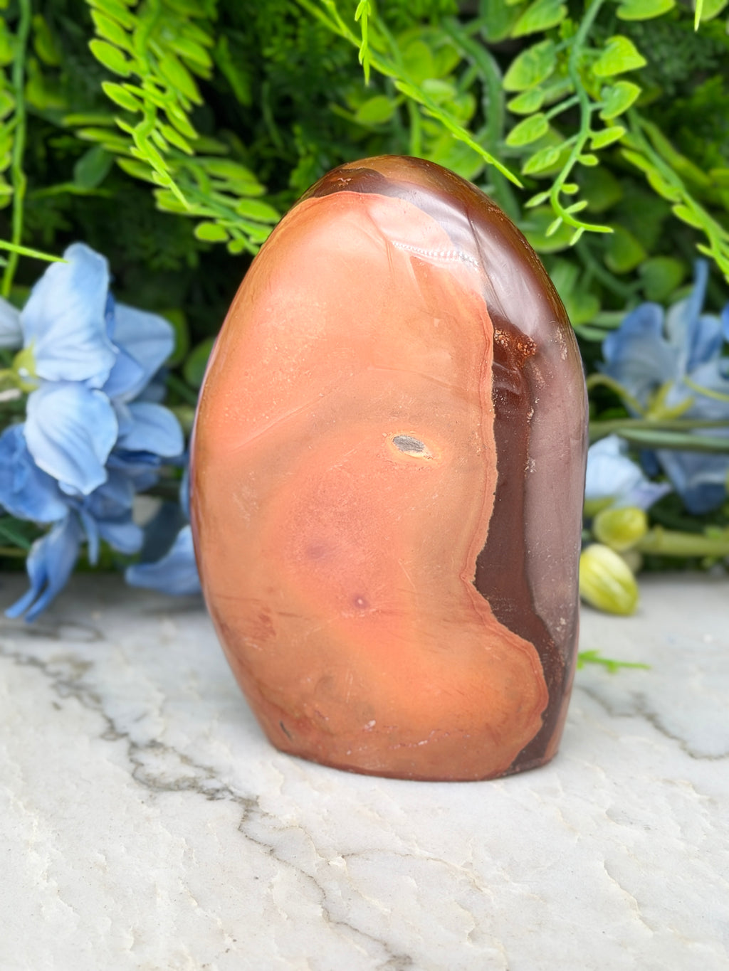 a brown and orange rock formation with a small hole in the center, surrounded by lush green foliage and blue flowers.