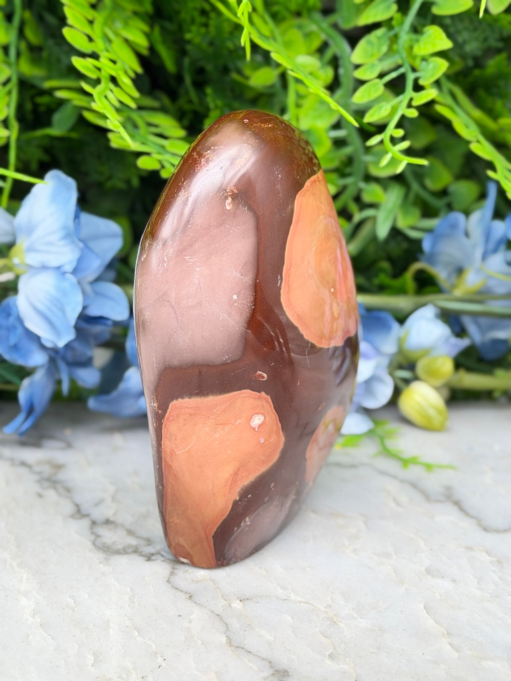 A brown and orange jadeite gemstone is displayed on a white marble surface, surrounded by blue flowers.