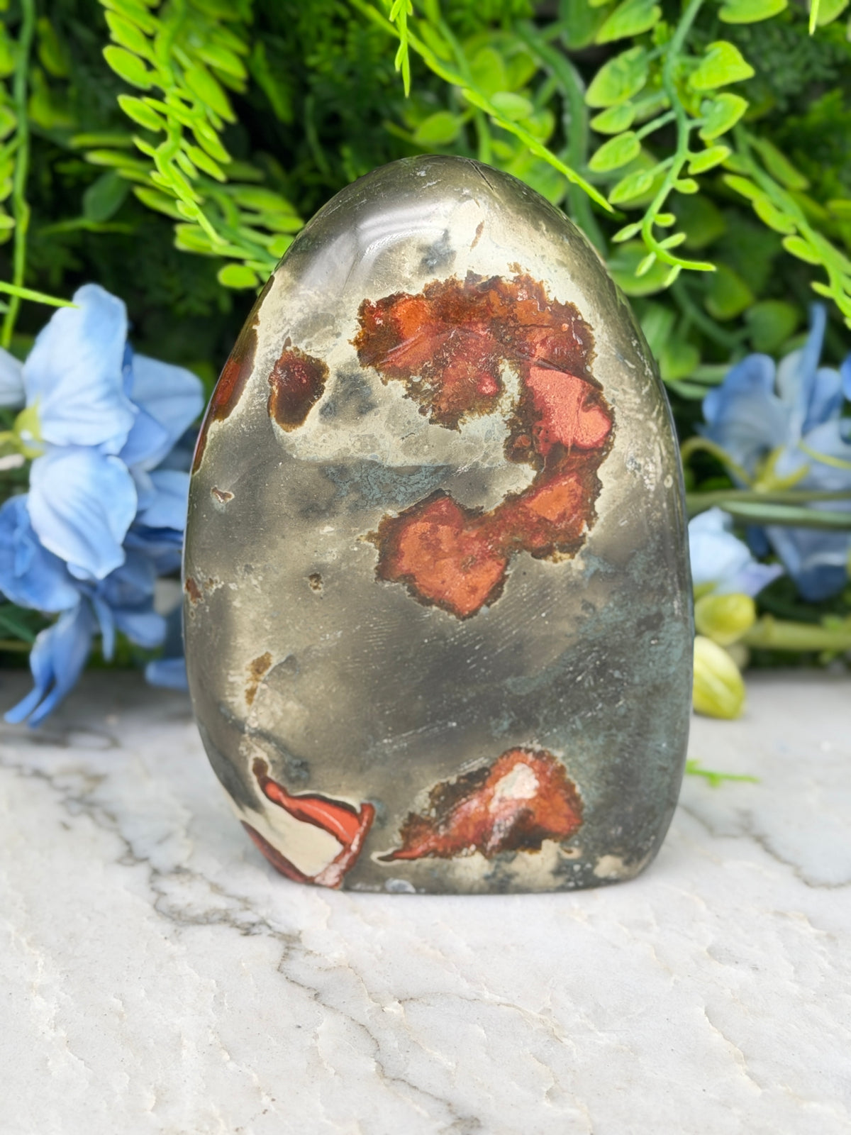 a jadeite gemstone with a unique, colorful pattern of red and orange hues, set against a backdrop of lush green foliage and blue flowers.