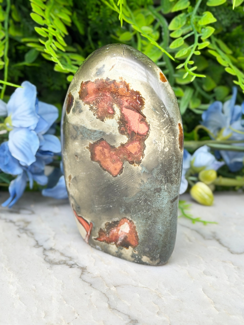 a large, irregularly shaped stone with a unique pattern of red and orange spots, surrounded by lush green foliage and blue flowers.