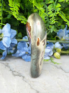 a unique, curved, and textured stone or metal object, possibly a piece of jewelry or a decorative item, placed on a white surface. The object has a distinctive shape and appears to be made of a material that resembles metal or stone. In the background, there are blue flowers, adding a touch of color and natural beauty to the scene.