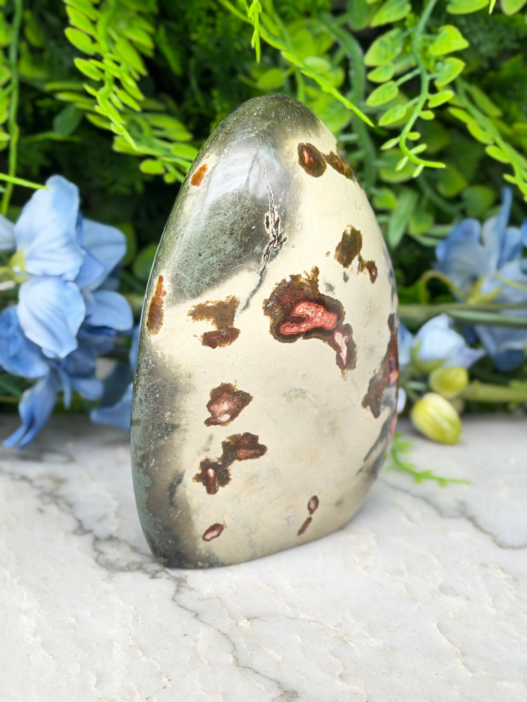 a jadeite gemstone with a speckled surface, surrounded by a lush green plant and blue flowers.