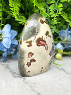 a jadeite gemstone with a speckled surface, surrounded by a lush green plant and blue flowers.