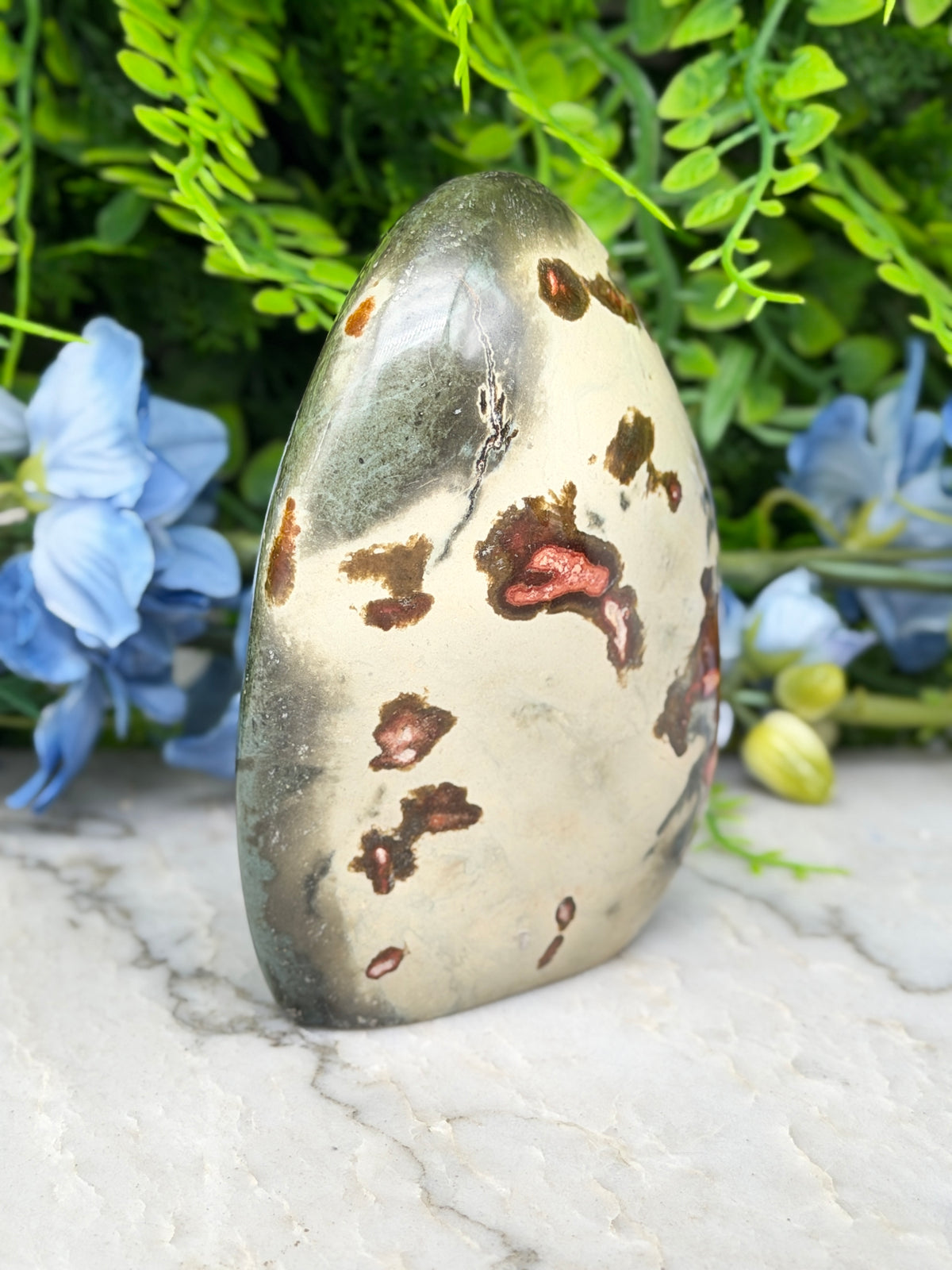 a jadeite gemstone with a speckled surface, surrounded by a lush green plant and blue flowers.