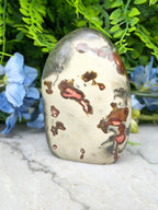 a jadeite crystal with a speckled surface, set against a backdrop of lush green foliage and blue flowers.