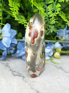 a jadeite gemstone with a unique, speckled pattern of red and brown hues, set against a backdrop of lush green foliage and delicate blue flowers.