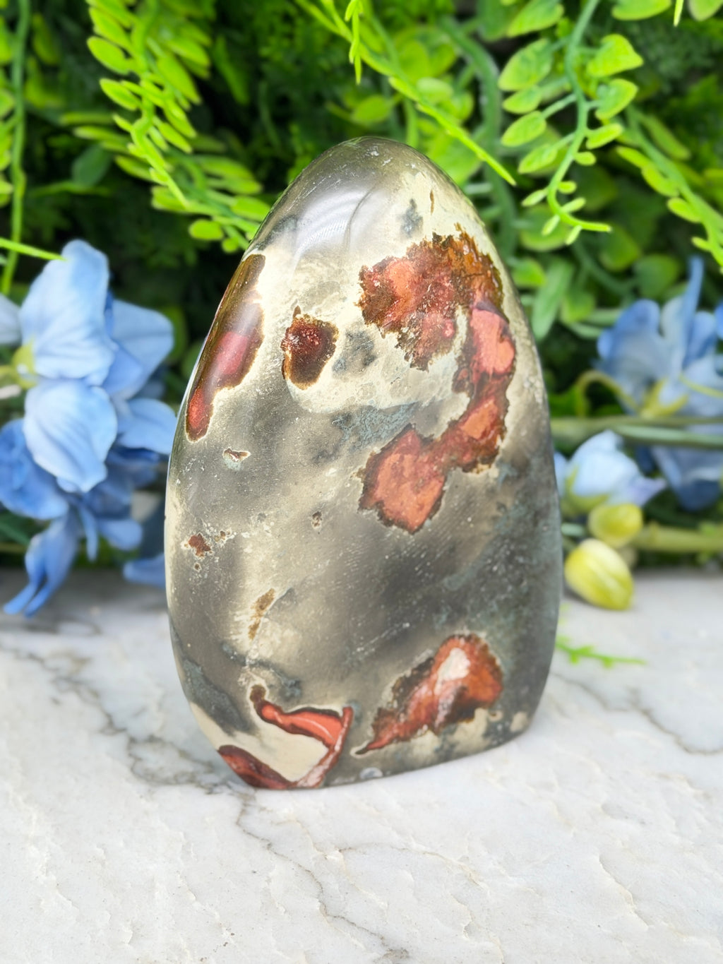 a jadeite gemstone with a unique, speckled pattern of red and brown hues, set against a backdrop of lush green foliage and delicate blue flowers.