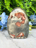 a jadeite gemstone with a unique, colorful pattern of red and orange hues, set against a backdrop of lush green foliage and blue flowers.