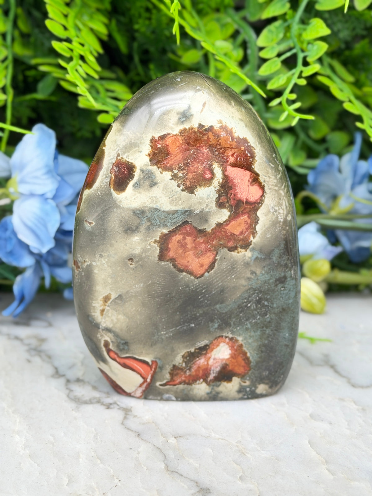 a jadeite gemstone with a unique, colorful pattern of red and orange hues, set against a backdrop of lush green foliage and blue flowers.