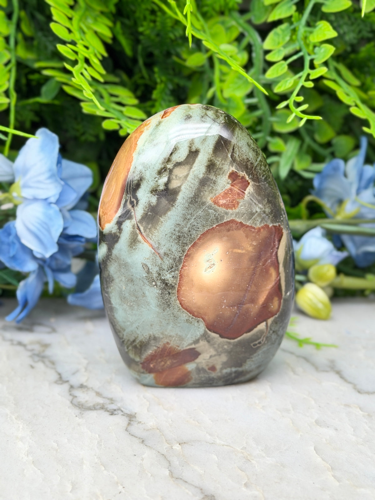 A jadeite, a type of jadeite gemstone, is displayed on a marble surface surrounded by blue flowers and greenery.
