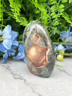 a large, irregularly shaped rock with a brown and green color pattern, placed on a white surface. The rock is surrounded by blue flowers, creating a visually appealing contrast.
