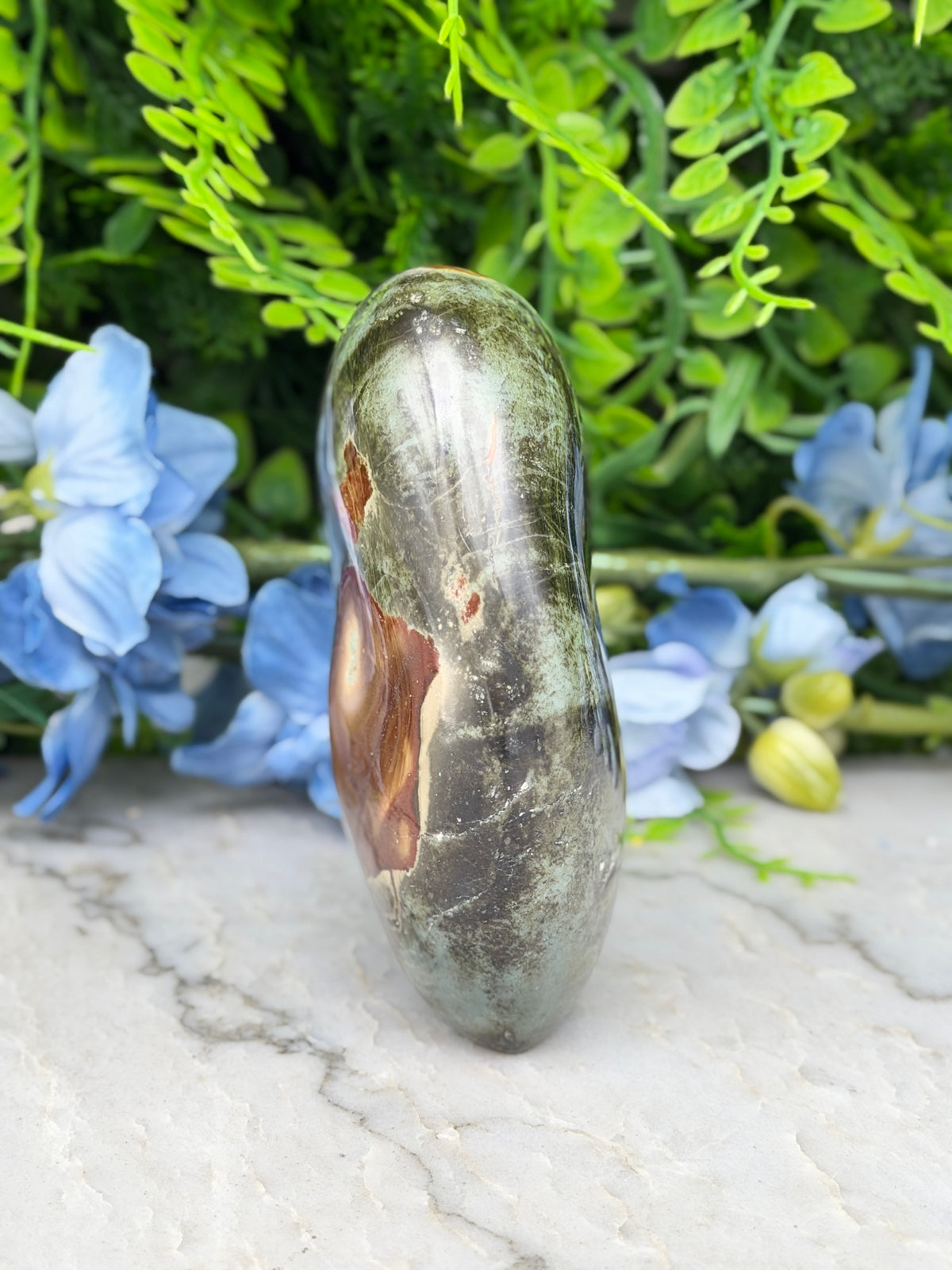 a large, dark green rock with a brownish hue, placed on a white surface. The rock is surrounded by a variety of blue and green flowers, creating a visually appealing contrast.