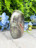 a jadeite, a type of natural stone, with a unique greenish-brown hue and a striking red stripe running through it, set against a backdrop of lush greenery and blue flowers.