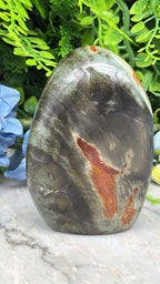 A close-up image of a jadeite gemstone, featuring a mix of black, red, and orange hues, set against a backdrop of lush green foliage.