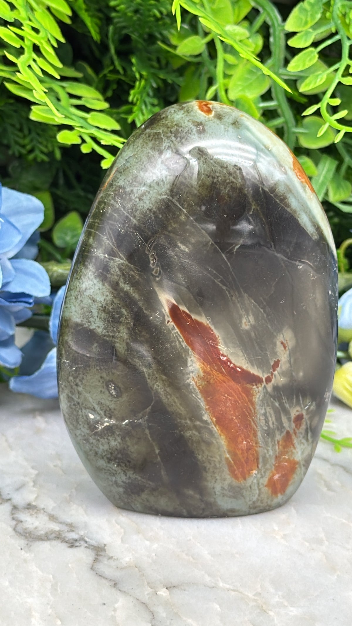 A close-up image of a jadeite gemstone, featuring a mix of black, red, and orange hues, set against a backdrop of lush green foliage.