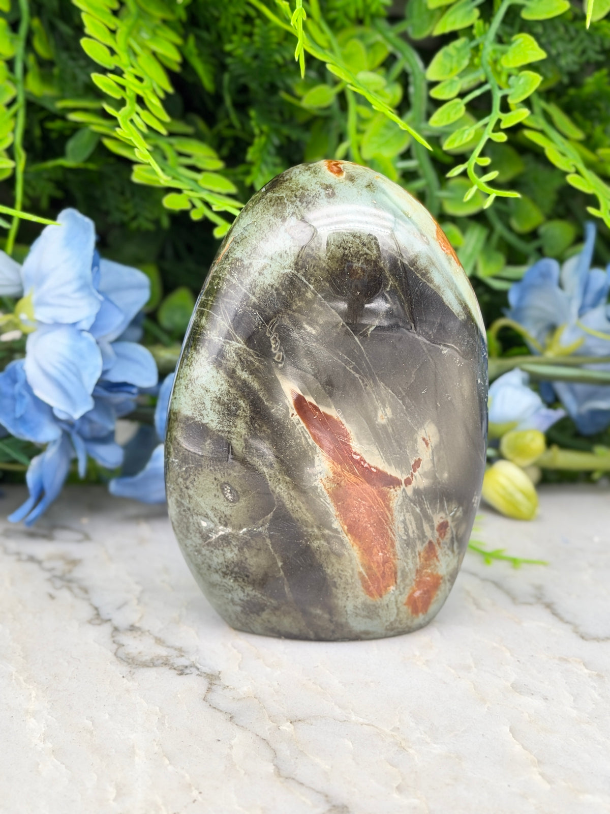 a jade stone with a unique, multicolored pattern, surrounded by lush green foliage and blue flowers.