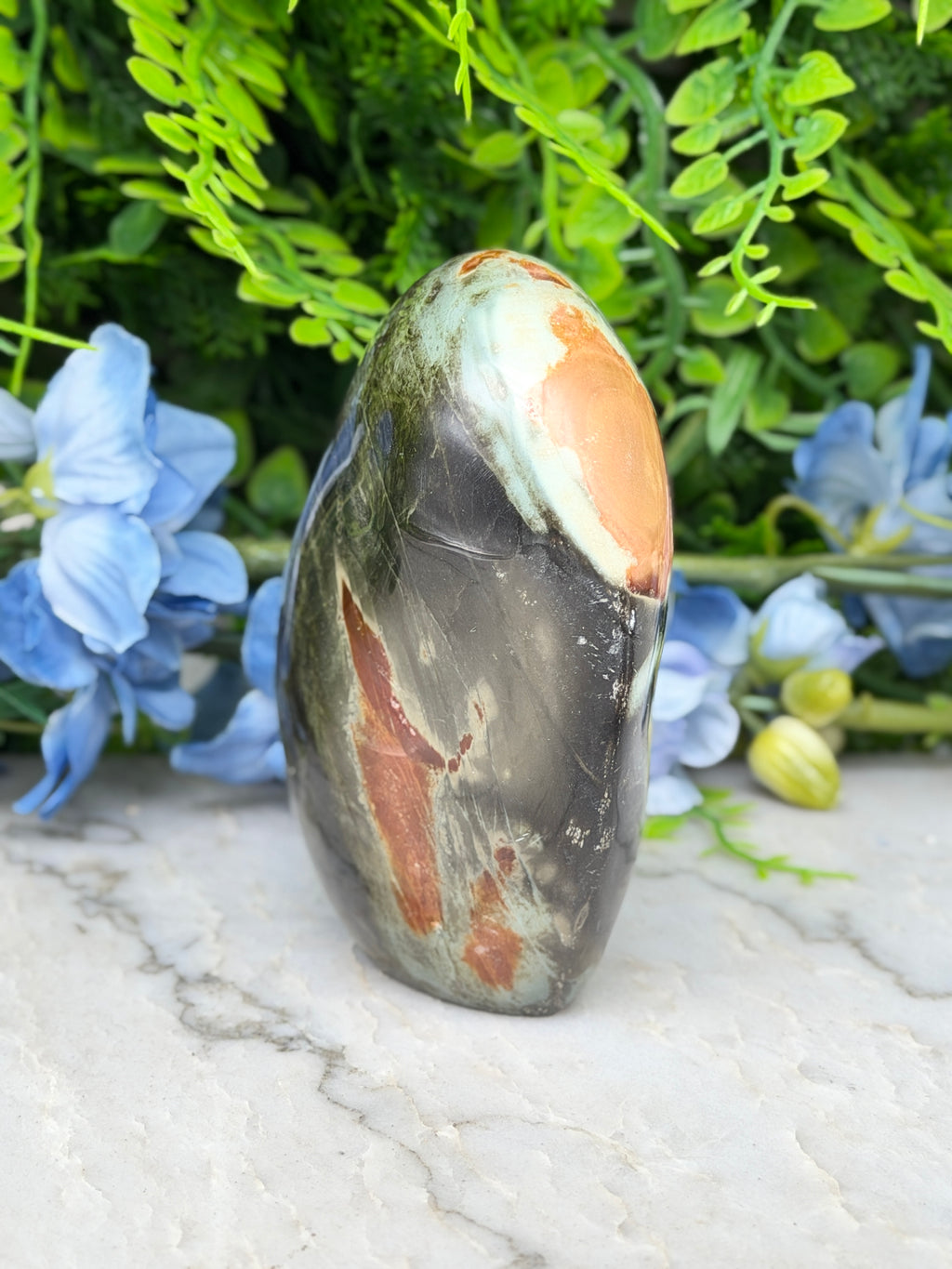 a large, multi-colored rock with a unique pattern, surrounded by blue flowers and green foliage.
