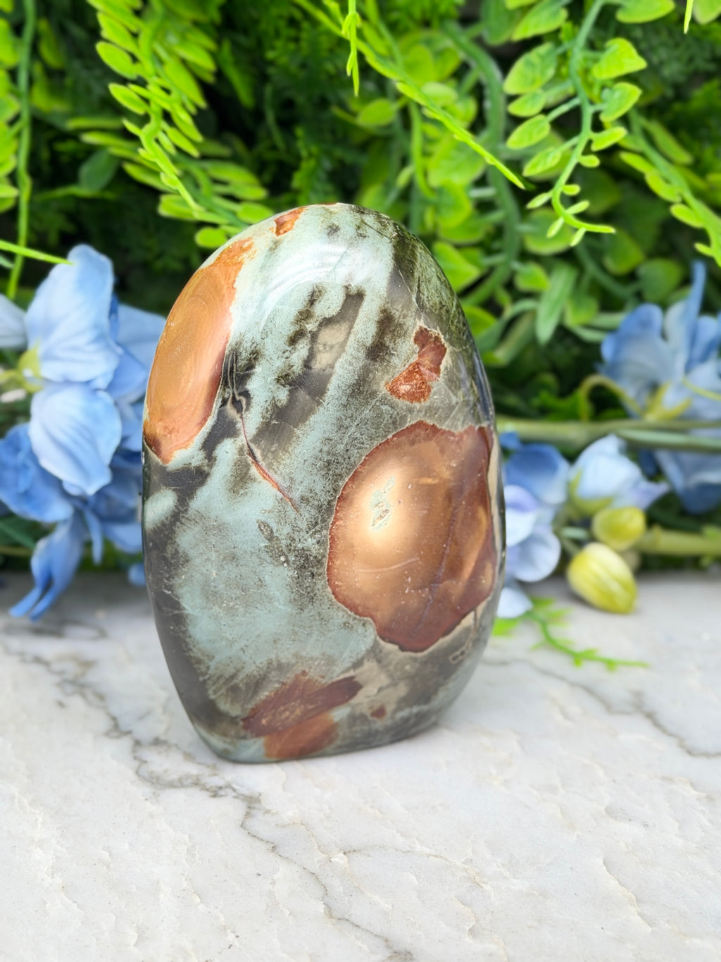 a large, irregularly shaped stone with a mix of green, brown, and orange colors, set against a backdrop of lush greenery and blue flowers.