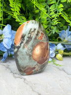 a large, irregularly shaped stone with a mix of green, brown, and orange colors, set against a backdrop of lush greenery and blue flowers.