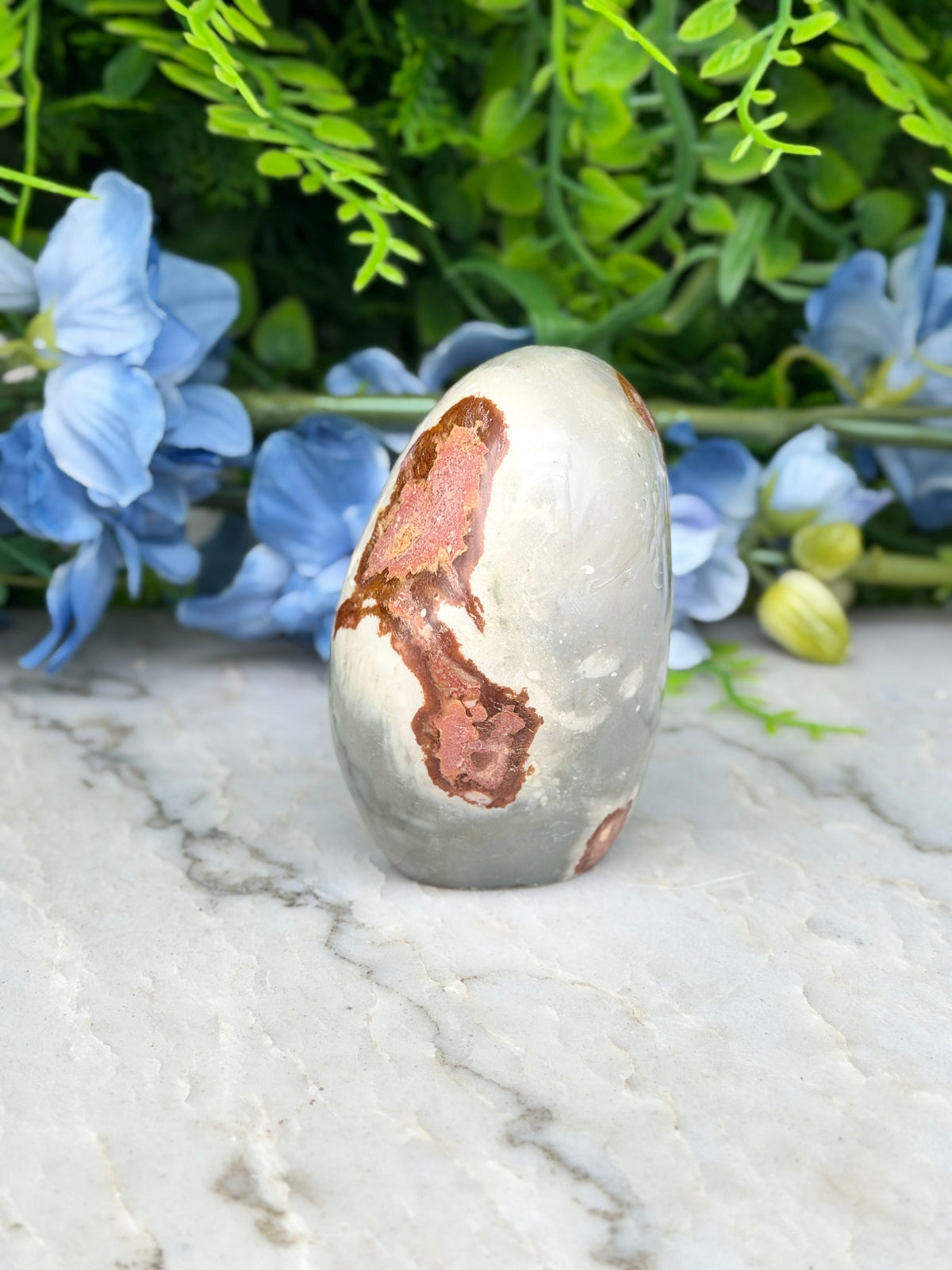 a white and brown rock with a unique pattern, placed on a marble surface. The rock is surrounded by blue flowers, creating a visually appealing contrast.